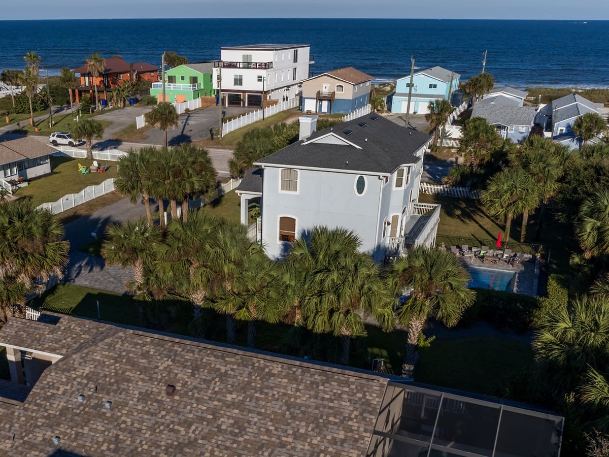 Aerial view of a two-storey coastal home with a private pool among palm trees, the beach houses of Vilano Beach and the Atlantic Ocean beyond it